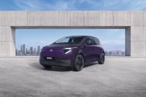 Smart, sjov og perfekt til byen: Den helt nye elektriske Nissan PIXO er her Three-quarter front view of a purple Nissan PIXO electric city car parked beneath a modern concrete structure, with a city skyline in the background.